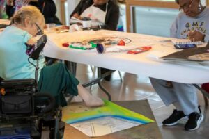 This image is of an older adult woman who is physically disabled in a wheelchair painting a picture on the ground with her feet. She has short blonde hair and glasses. She is wearing a light blue shirt.