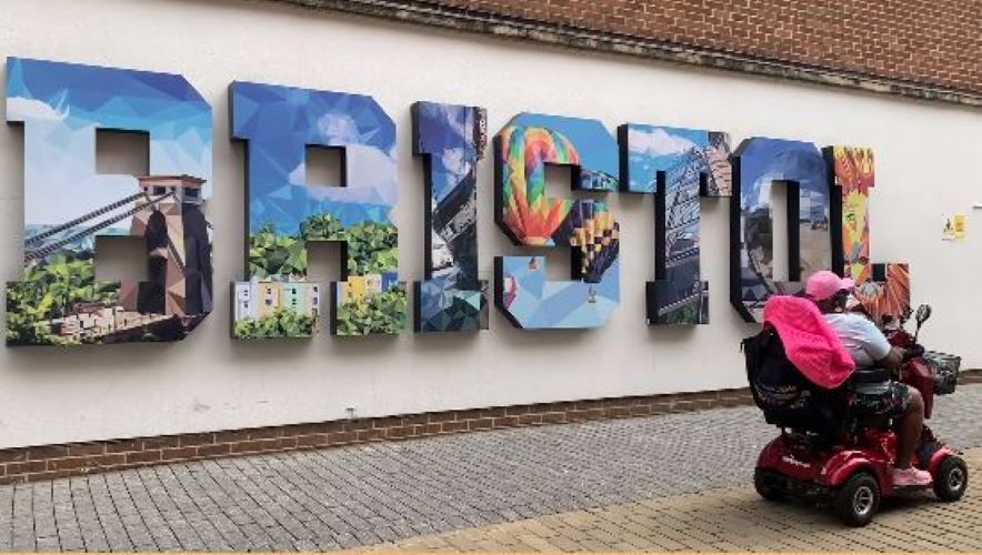 An older adult woman rides a motorized scooter with a large mural behind her that says, "Bristol."