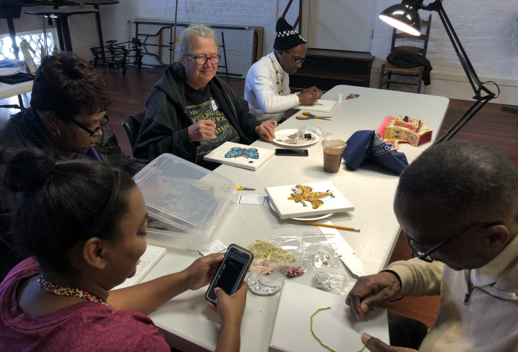 A group of four older adults, black and white, sit at a table and work on beading projects with teaching artist, Big Chief Darryl Montana, at the Louisiana State Museum in New Orleans